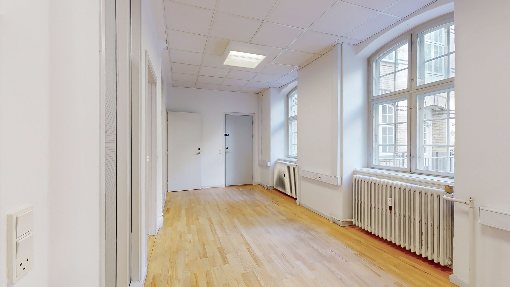 Kort korridor med lysegul gulvbelægning og hvide vægge og radiatorer. Højt loft med indbygget lys og store ruder med udsigt