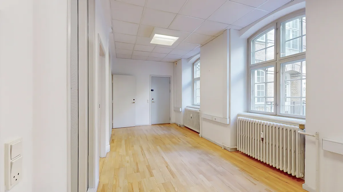 Kort korridor med lysegul gulvbelægning og hvide vægge og radiatorer. Højt loft med indbygget lys og store ruder med udsigt