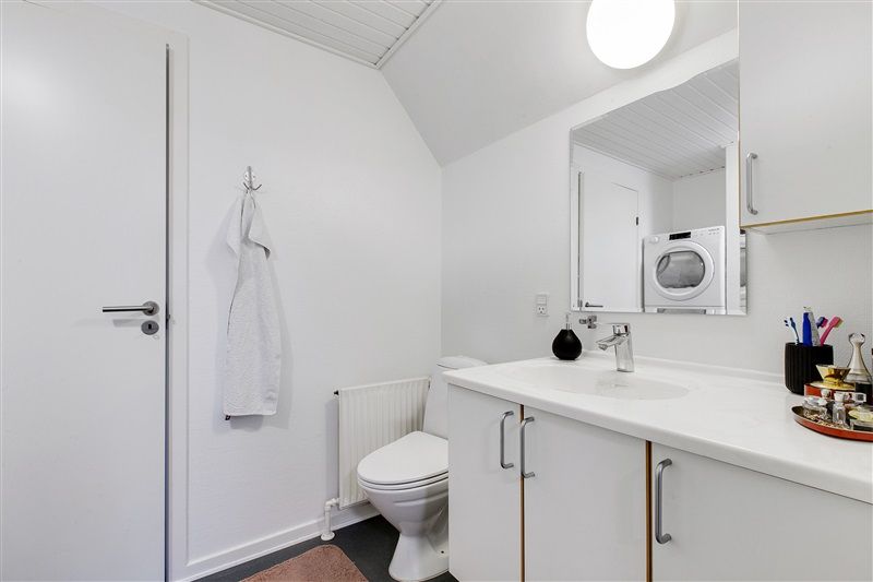 Hvidt badeværelse med hvide skabe og toilet samt hvidt vasketøjstøjle og spejl med toiletpapir og sæbeflaske på skabet