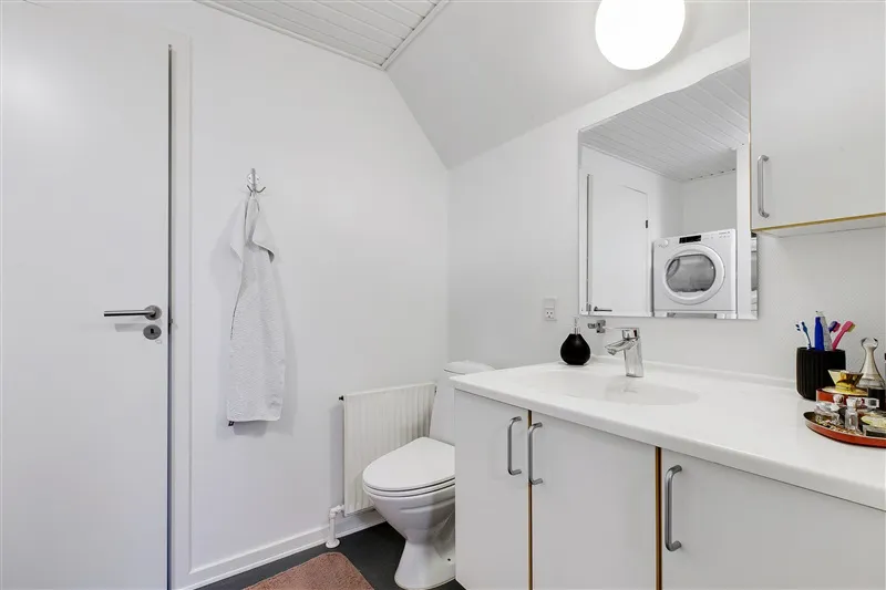 Hvidt badeværelse med hvide skabe og toilet samt hvidt vasketøjstøjle og spejl med toiletpapir og sæbeflaske på skabet