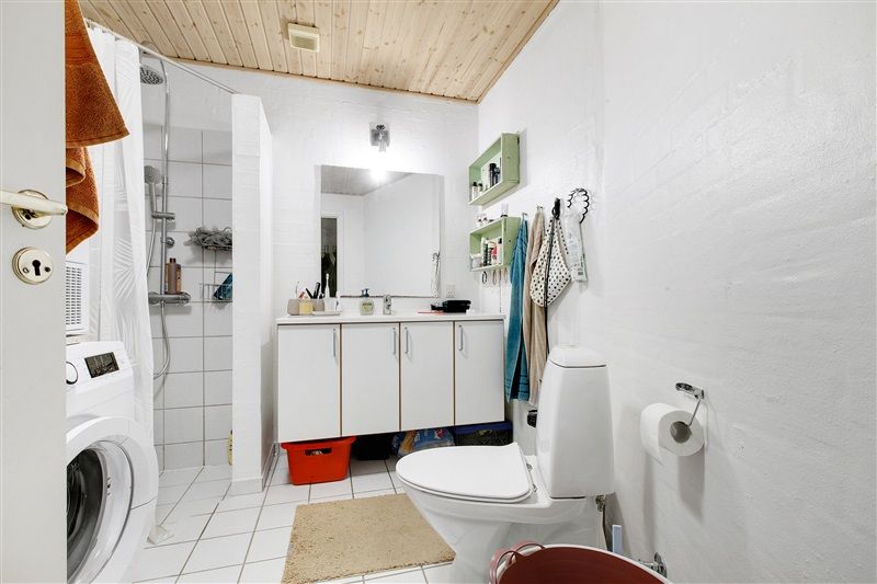 Et moderne badeværelse med hvide vægge, en hvid toilet, en vask med spejl, hvide reoler og en hvid tøjvask