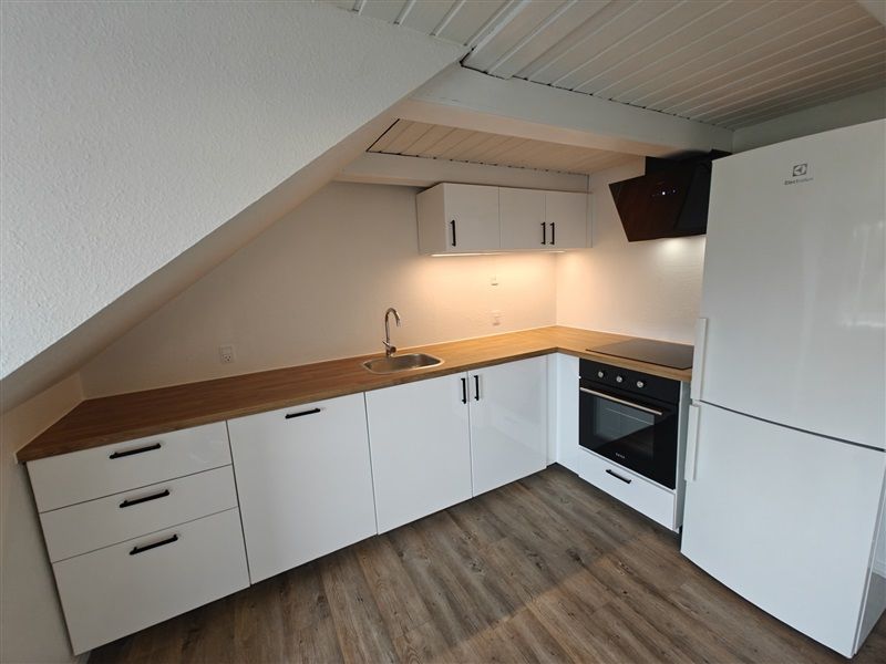 En velholdt køkken med hvide skabe, lysegule trægulv, køleskab og komfur samt et enkelt hængelampe over øen