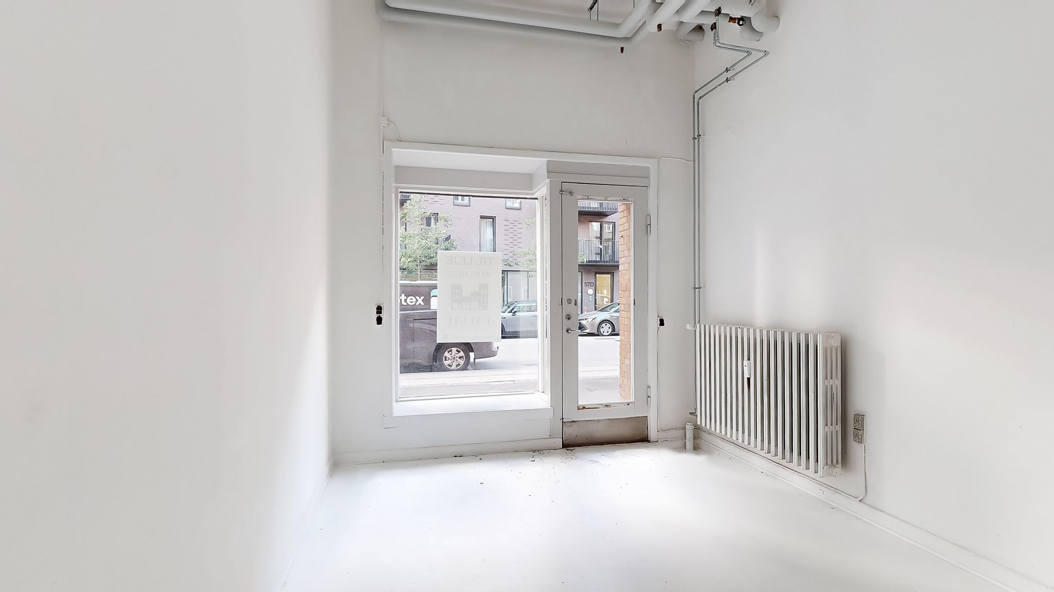 En lejlighedsdør og et vindue er åbne mod en parkeringsplads med biler og et hus med altan