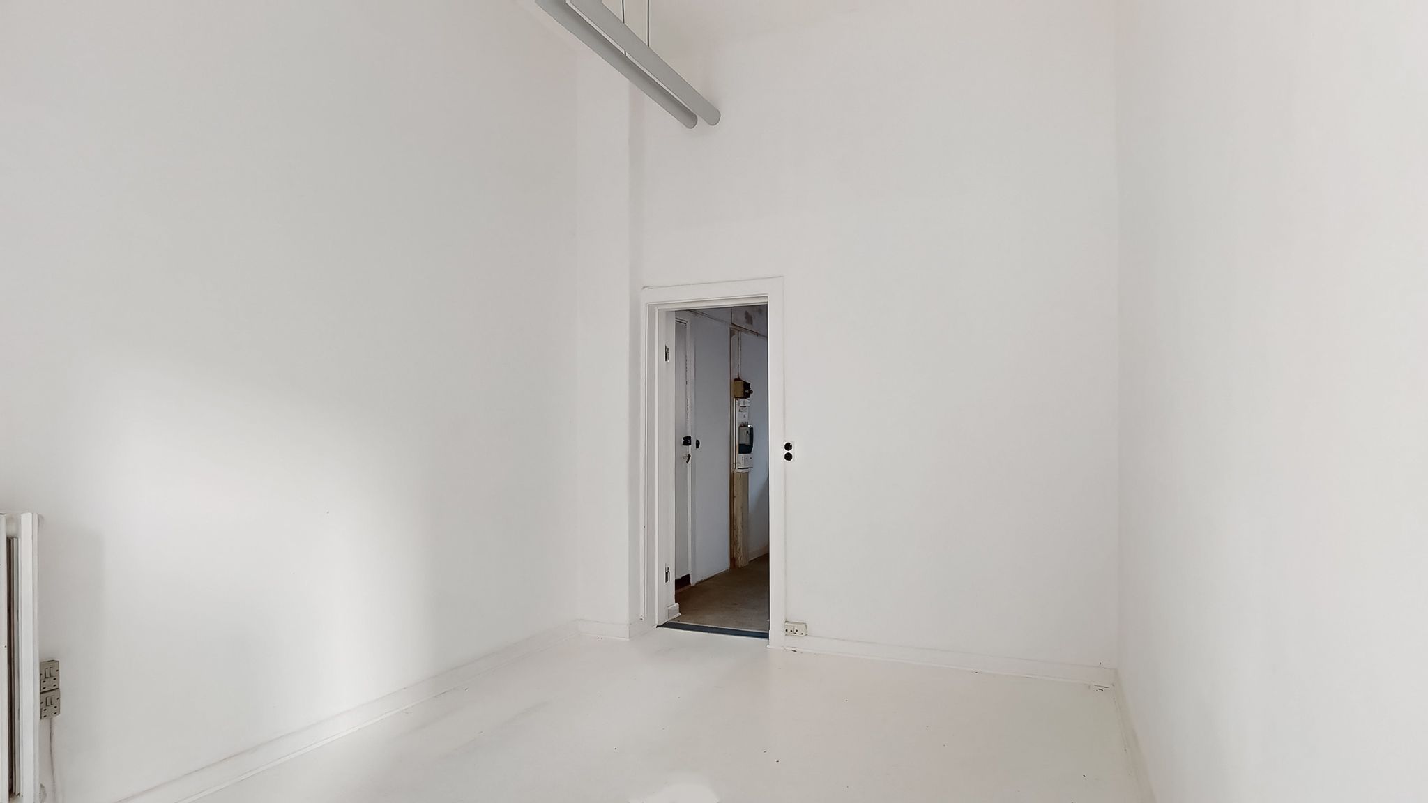 To hvide vægge, hvor en dør er åben, og der er en lampe, der hænger fra loftet, samt en radiator og en stikkontakt på højre