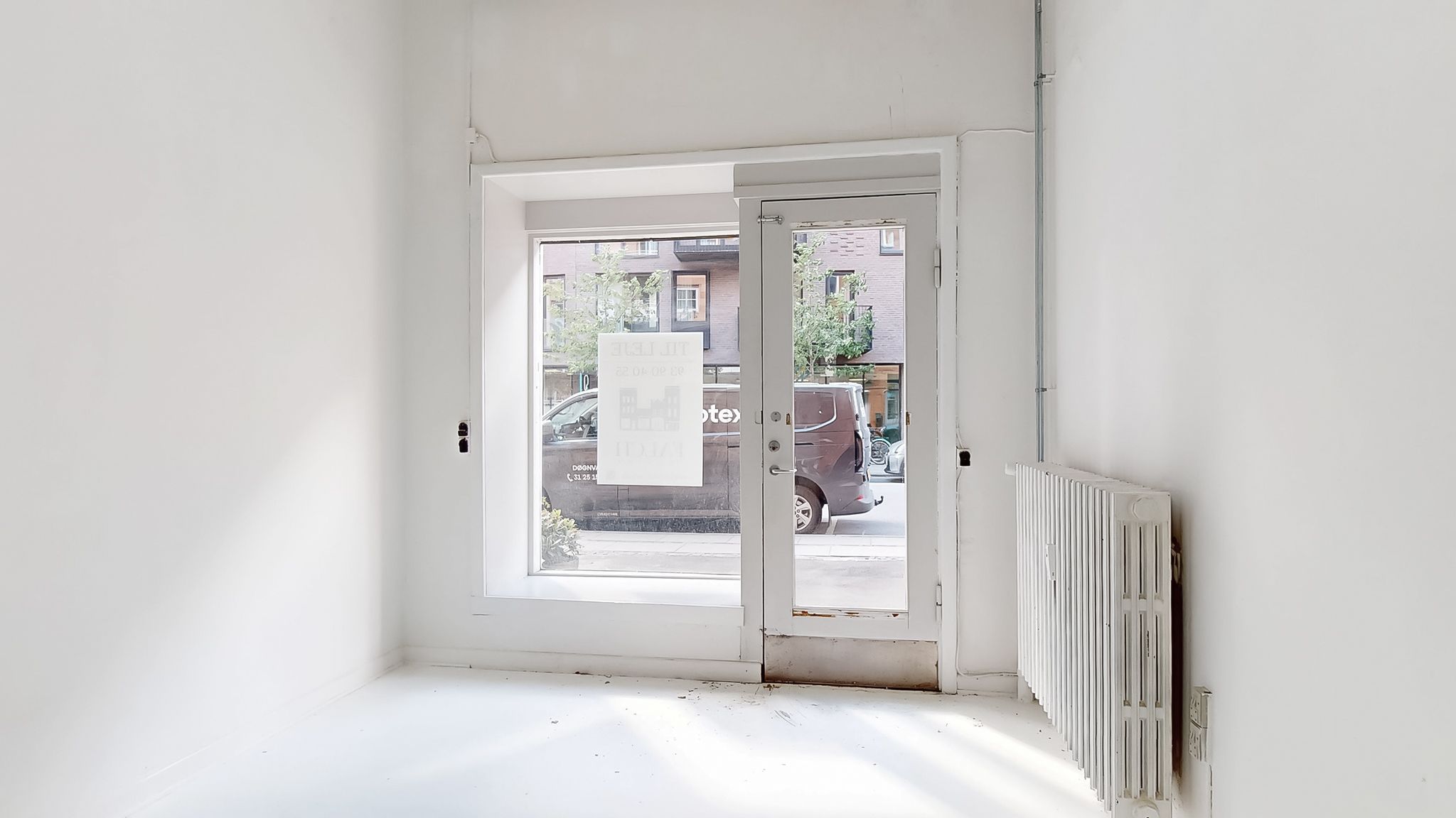 En tom stue med hvide vægge og gulv samt glasdør med annonceskiltet "Leje lejlighed" og et hvidt radiator til højre