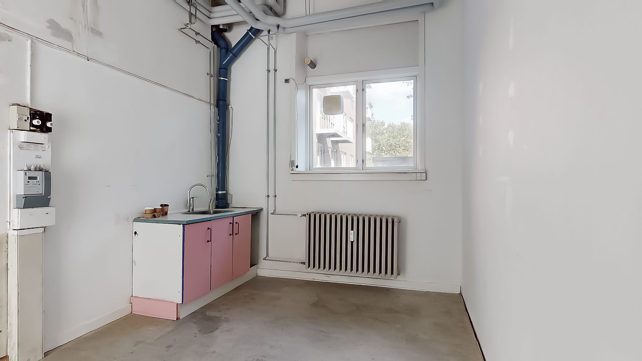 En tom stue med hvide vægge, loftsrør og radiatorer, samt et køkken med pink og hvide skuffer, hvor der er en vandhane og