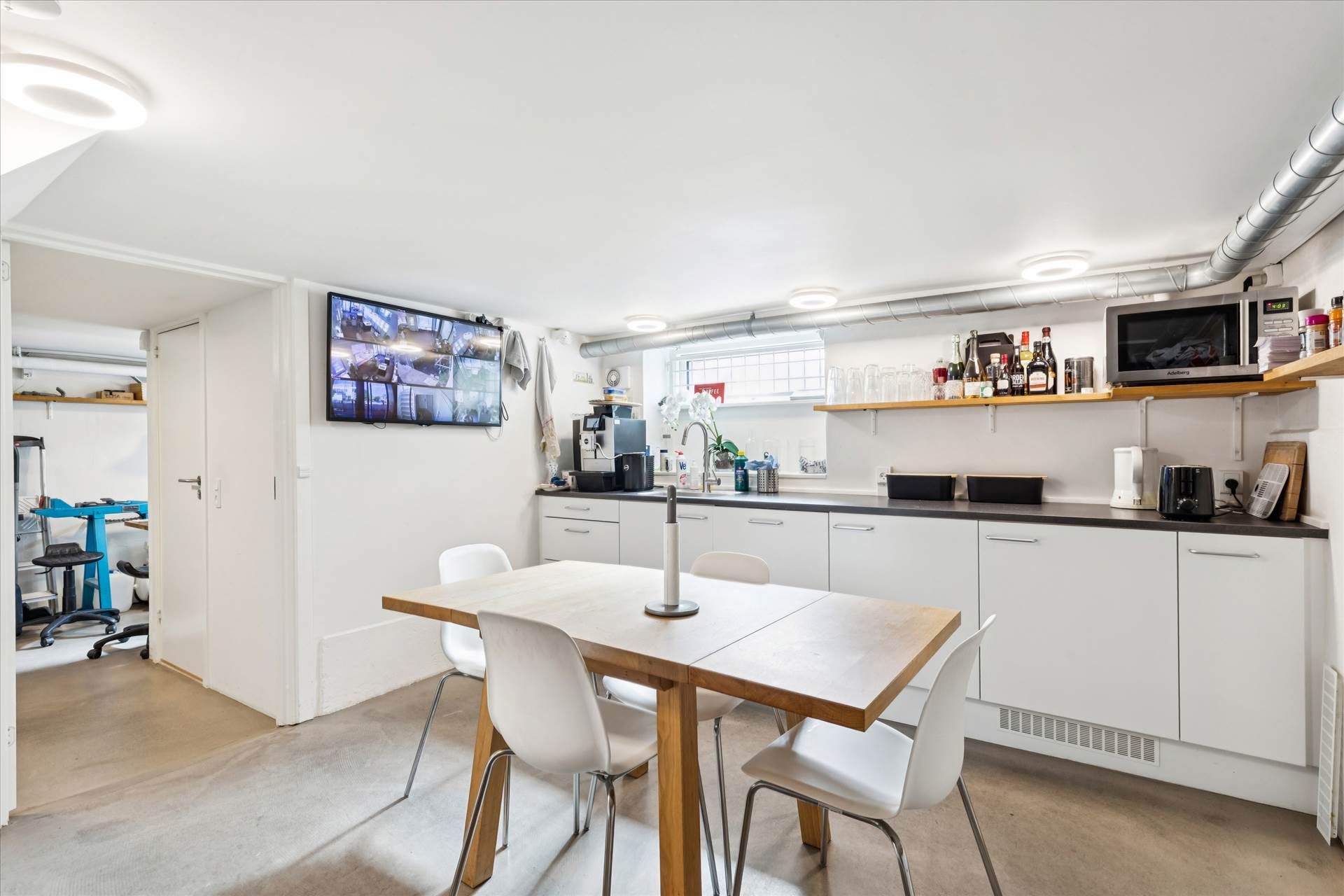 Et køkken med hvidt bord og stole, køkkenapparater og en tv-skærm med kameraoptagelser.