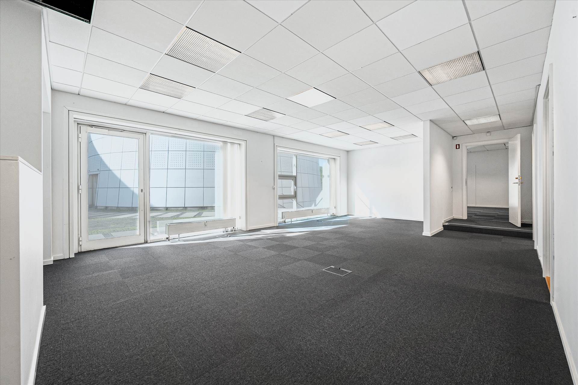 Et stort, lyse, tomt kontor med tæppegulv og hvide vægge samt store glasruder og vinduer