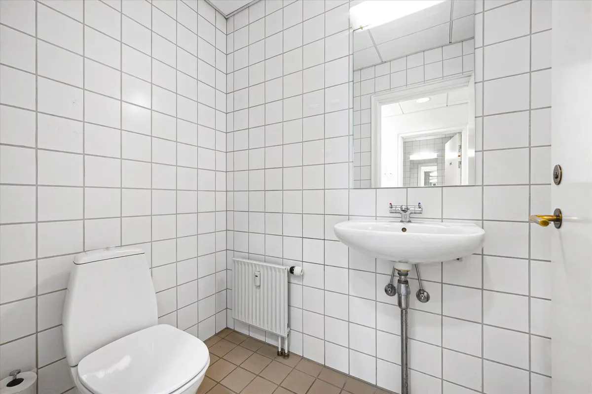 Et rent badeværelse med hvide flisede vægge, hvidt toilet og hvidt vask. En spejl over vasken og radiator ved siden af