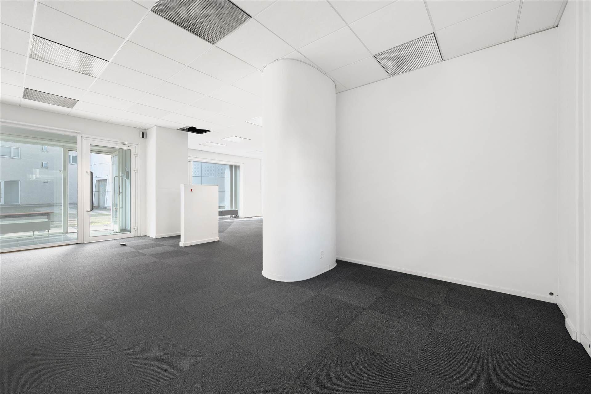 Kontorloft med hvide vægge, store vinduer, gulv med mørk gulvbelægning og en hvid søjle midt på gulvet
