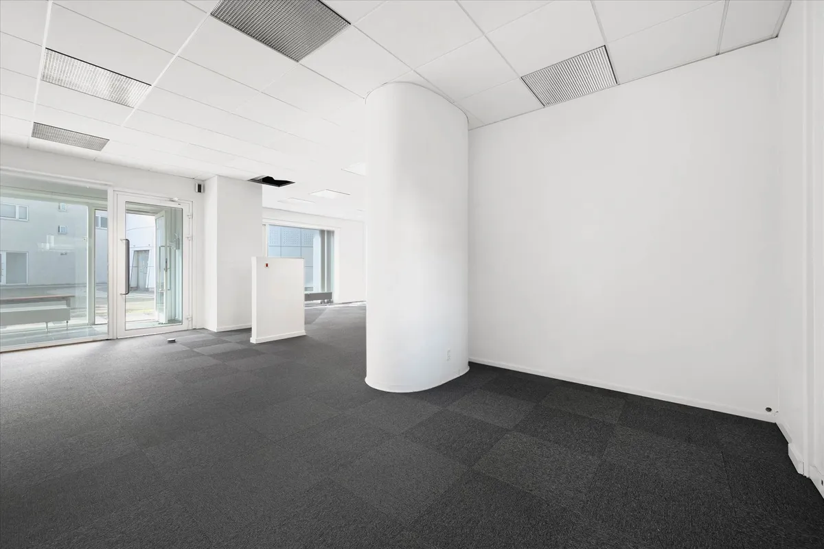 Kontorloft med hvide vægge, store vinduer, gulv med mørk gulvbelægning og en hvid søjle midt på gulvet