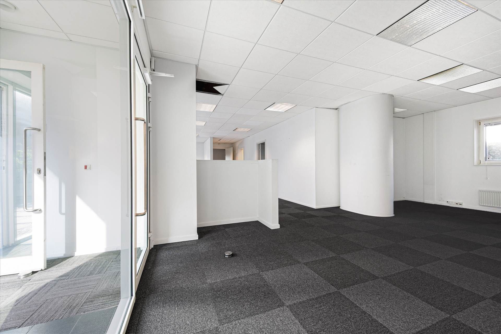 Kontorlokaler med hvide vægge, loft og gulv samt søjler og glasdøre med håndtag. Luftkanaler er installeret i loftet, og der
