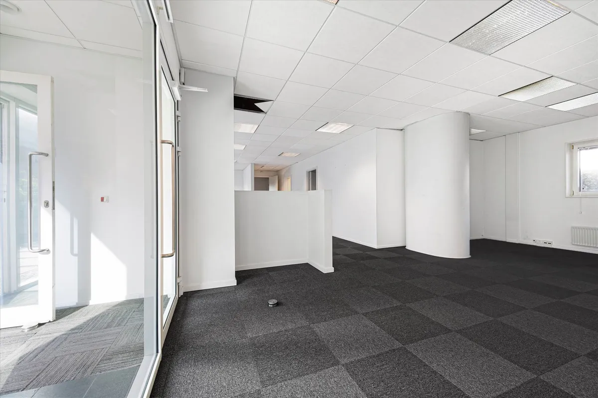 Kontorlokaler med hvide vægge, loft og gulv samt søjler og glasdøre med håndtag. Luftkanaler er installeret i loftet, og der