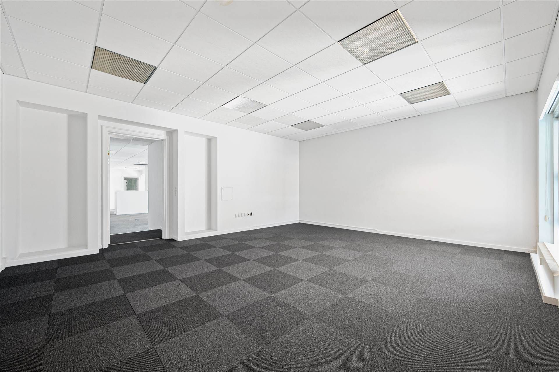 Rummelige kontorlokaler med hvide vægge, krydsfinerbelagt gulv og slanke loftspaneler. Et lukket dørstykke og en stor