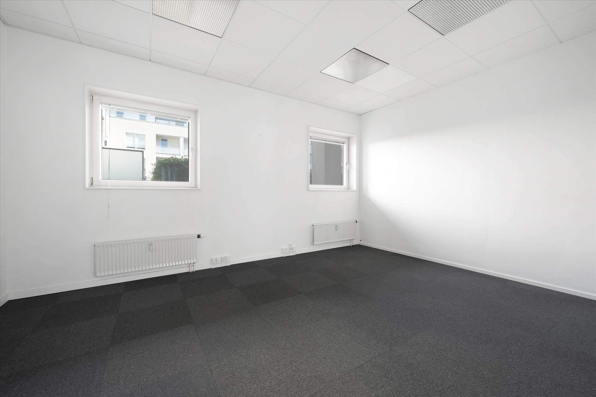 Kontorlokaler med to vinduer og to radiatorer, hvide vægge og loft samt gulvbelægning af gråt materiale.