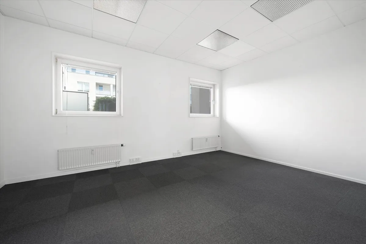 Kontorlokaler med to vinduer og to radiatorer, hvide vægge og loft samt gulvbelægning af gråt materiale.