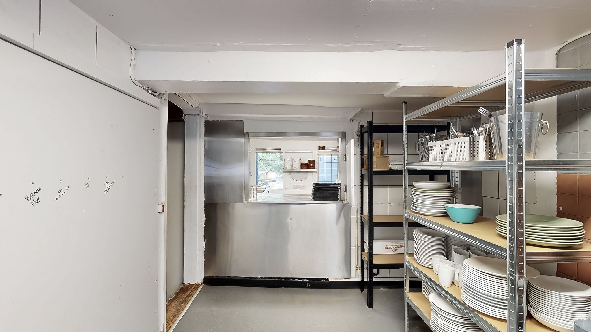 Et lager med mange hvide tallerkener, kopper og tallerkener, samt en metalhylde med køkkenredskaber og et køleskab med et