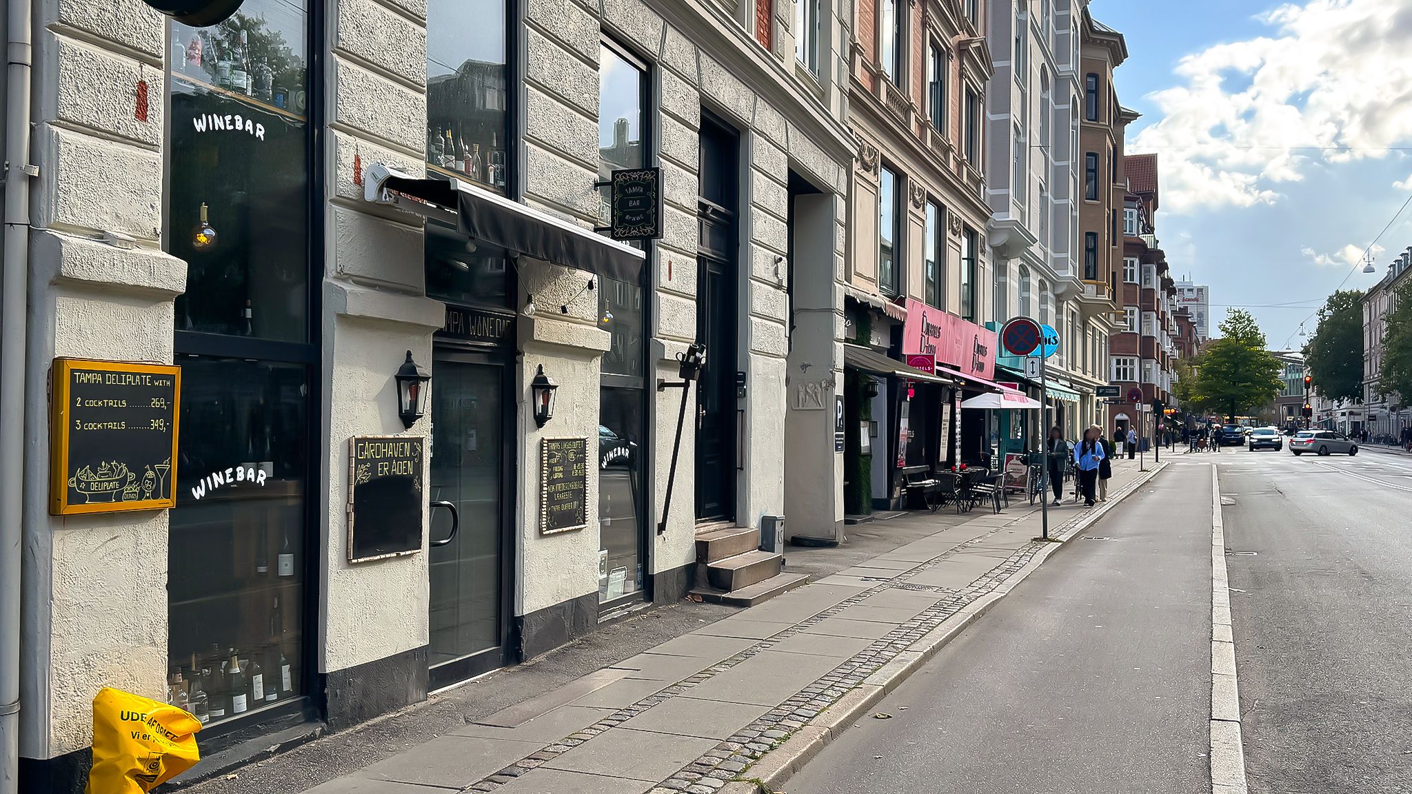 En tom gade med butikker og restauranter og folk, der går på fortovet. Et af lokalerne er en vinbar med skilte og menukort i