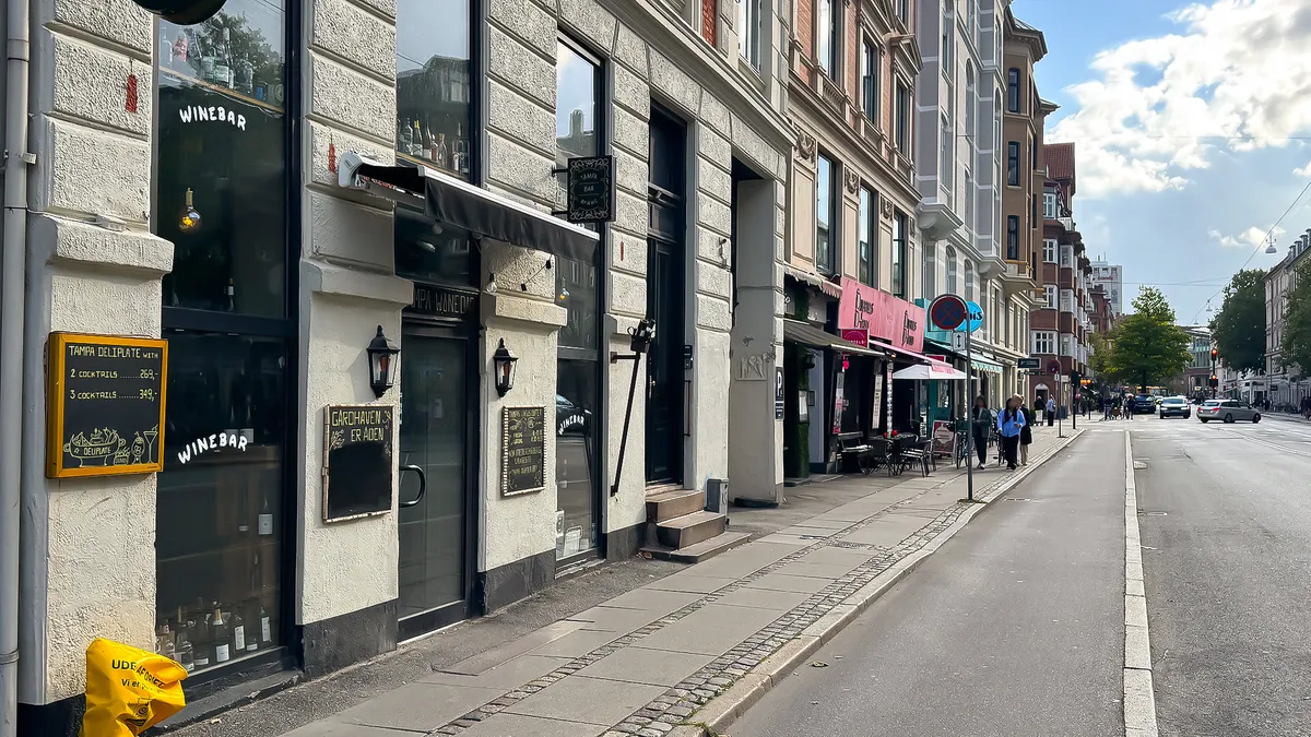 En tom gade med butikker og restauranter og folk, der går på fortovet. Et af lokalerne er en vinbar med skilte og menukort i