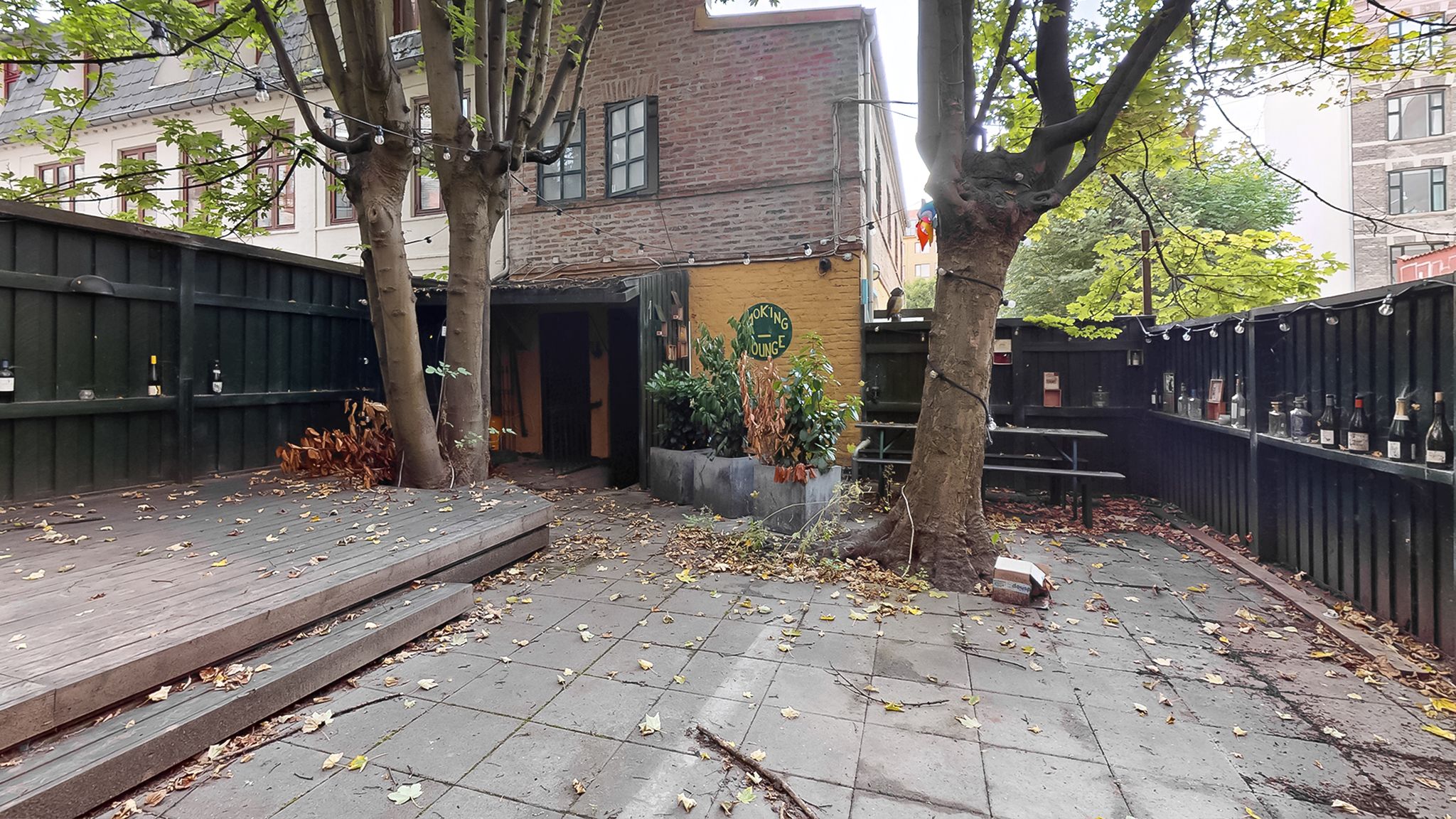 En gårdhave med en terrasse, søjler, planter og flasker af vin. På facaden står der "cooking lounge".