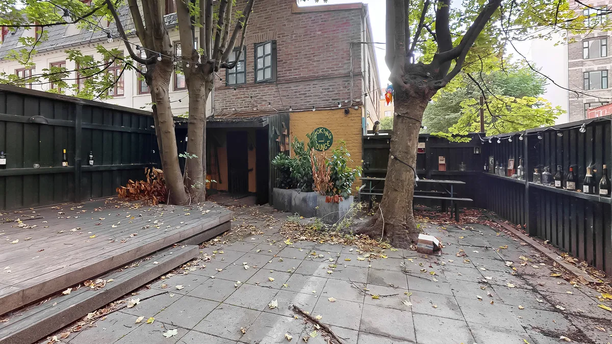 En gårdhave med en terrasse, søjler, planter og flasker af vin. På facaden står der "cooking lounge".