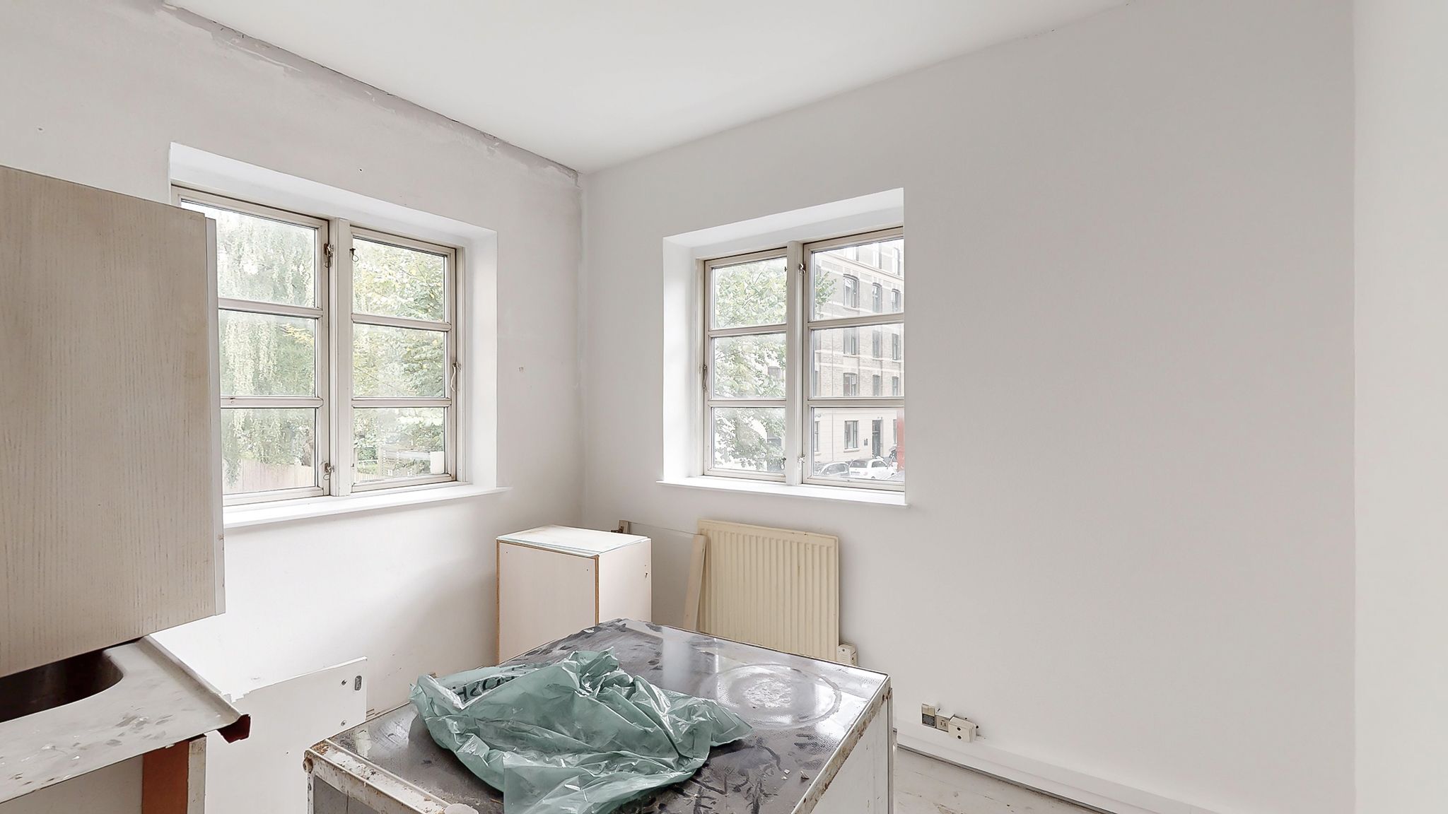 To tomme vægge i et lejlighedsværelse med to vinduer og en hvid radiator samt en køkkenbord og en plastpose