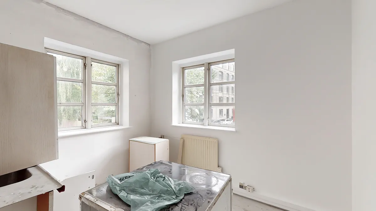 To tomme vægge i et lejlighedsværelse med to vinduer og en hvid radiator samt en køkkenbord og en plastpose