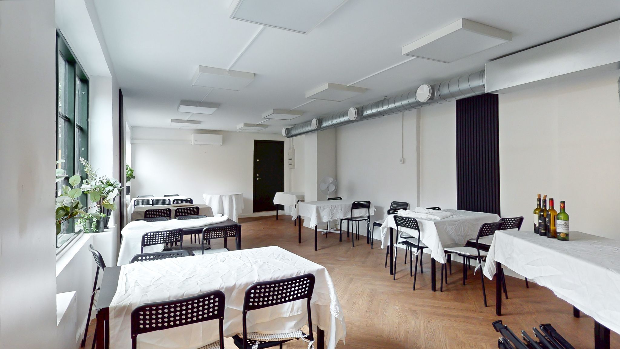En tom konferencelokale med hvide borde og stole, vinflasker på bordene og et klimaanlæg i væggen