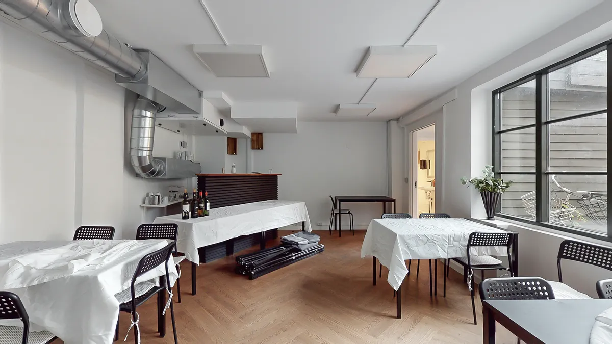 Inde i et hvidt lokale med trægulve og højt loft er der flere borde og stole. Der er en lang bar med vinflasker og en plante