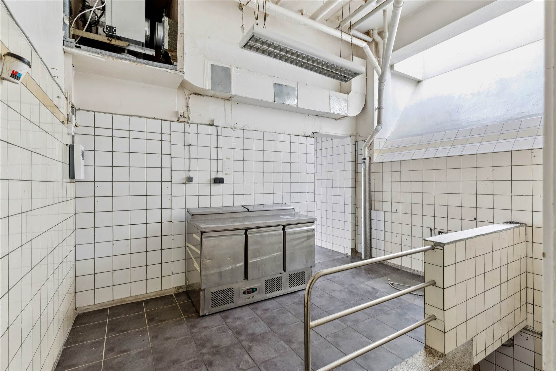 Køkken med højloft og metalkøleskab samt håndtag og hvidt klethedspanel i en kommerciel bygning