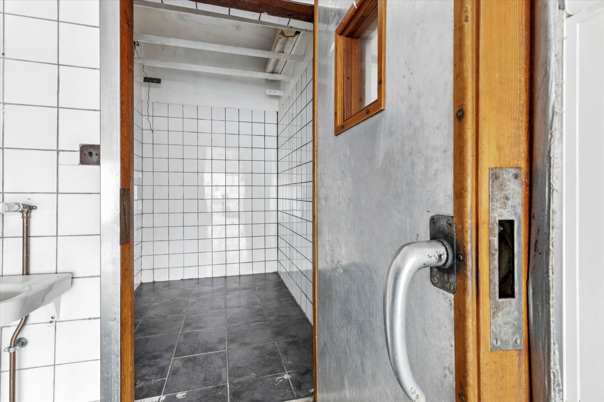 En åben dør til et lille badeværelse med hvide kakelvægge, sort gulv og en lille, firkantet rude i en træindfatning. Der er