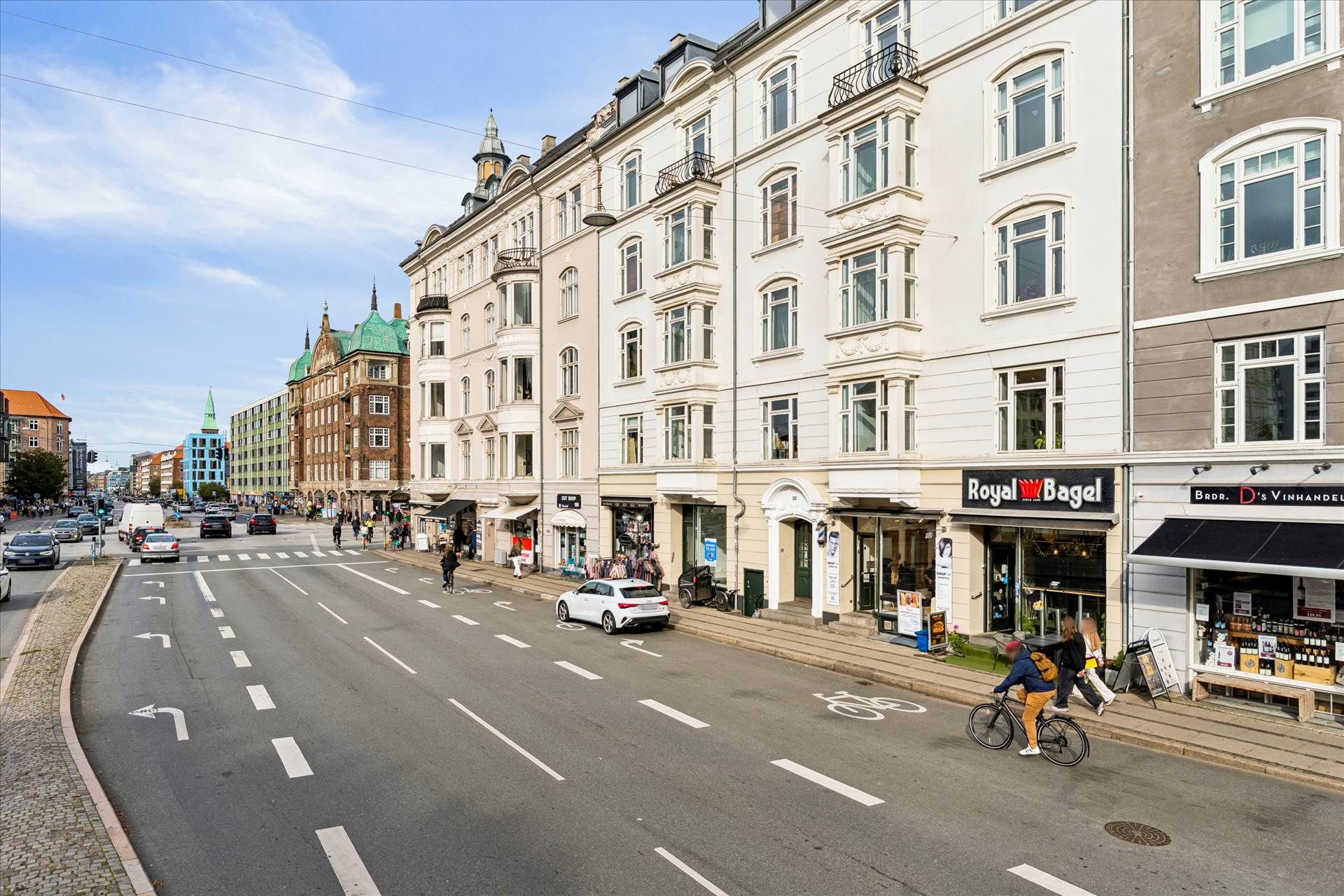 En bred gade med biler og cyklister, forretninger med Royal Bagel-skilt og tekst på dansk, mennesker og træer i baggrunden