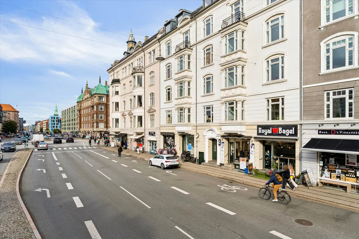 En bred gade med biler og cyklister, forretninger med Royal Bagel-skilt og tekst på dansk, mennesker og træer i baggrunden