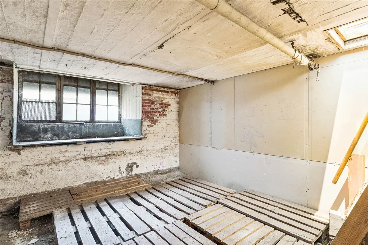 En gammel kælder med hulloft, vinduer og trægulve samt en trappe. Væggene er delvist afskallet og der er skraldespand og