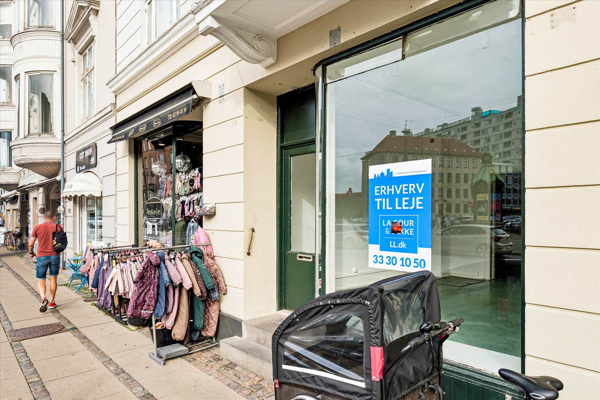 En mand går forbi en butik med en klapvogn parkeret foran indgangen og en reklame for en ny lejekontrakt på facaden