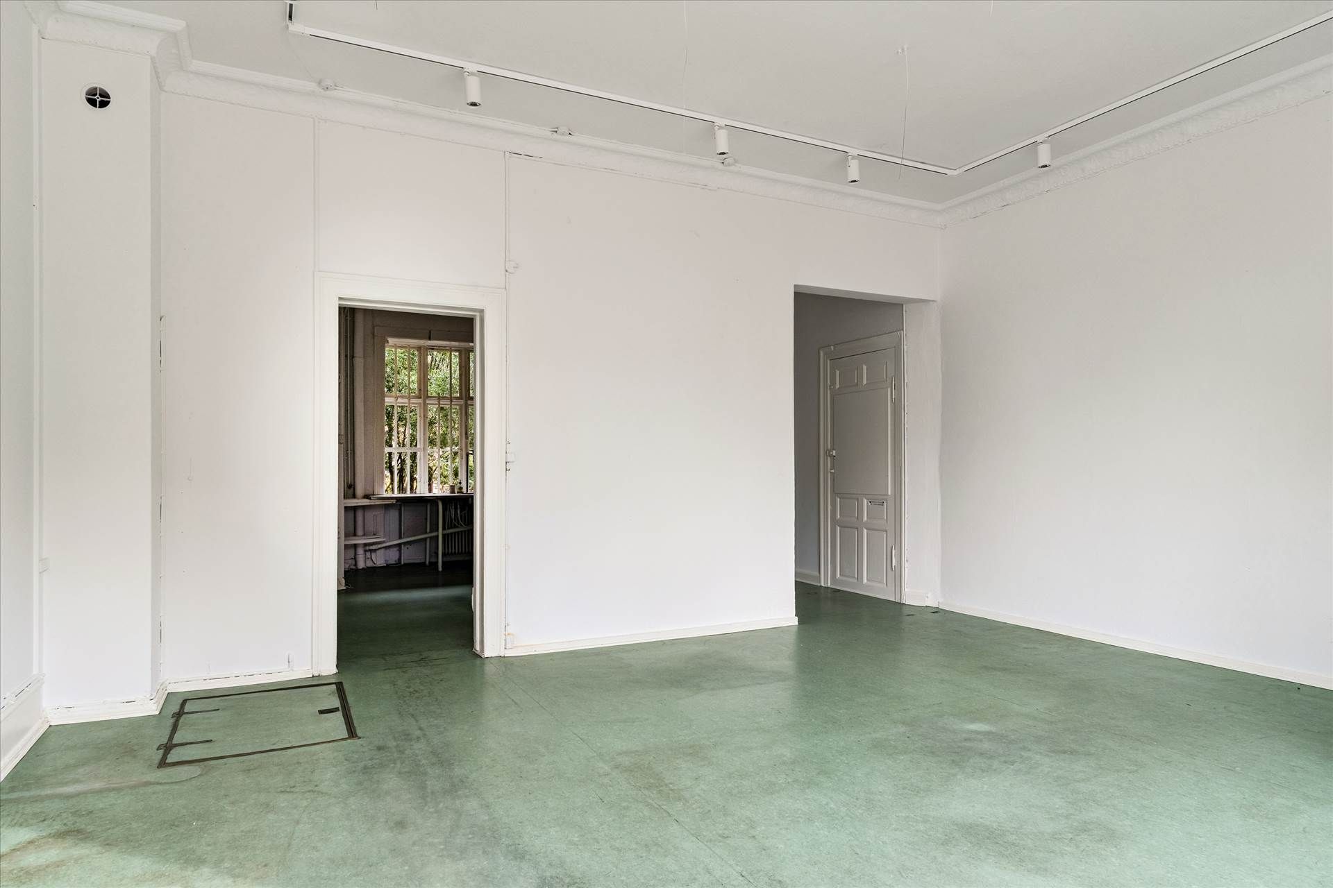 En stor, ren, hvid stue med grønne gulvbelægning og åbne døre ud mod en mindre stue og et mindre rum med vinduer