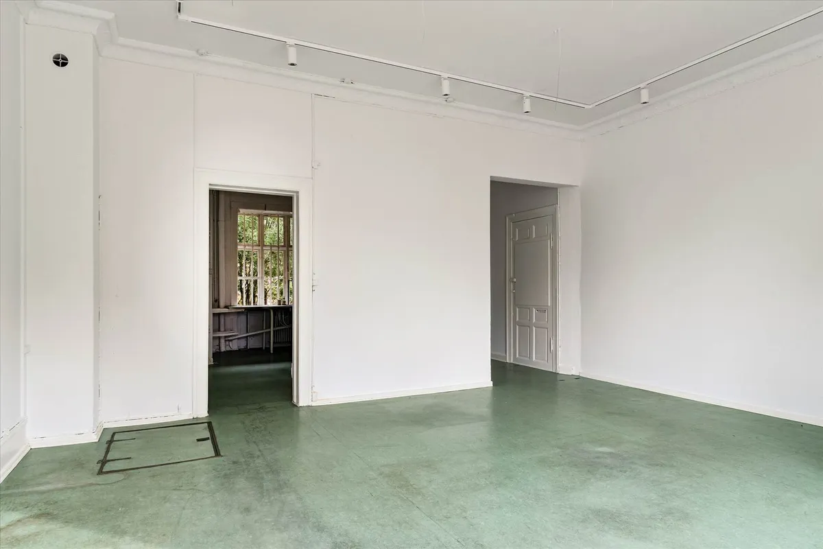 En stor, ren, hvid stue med grønne gulvbelægning og åbne døre ud mod en mindre stue og et mindre rum med vinduer