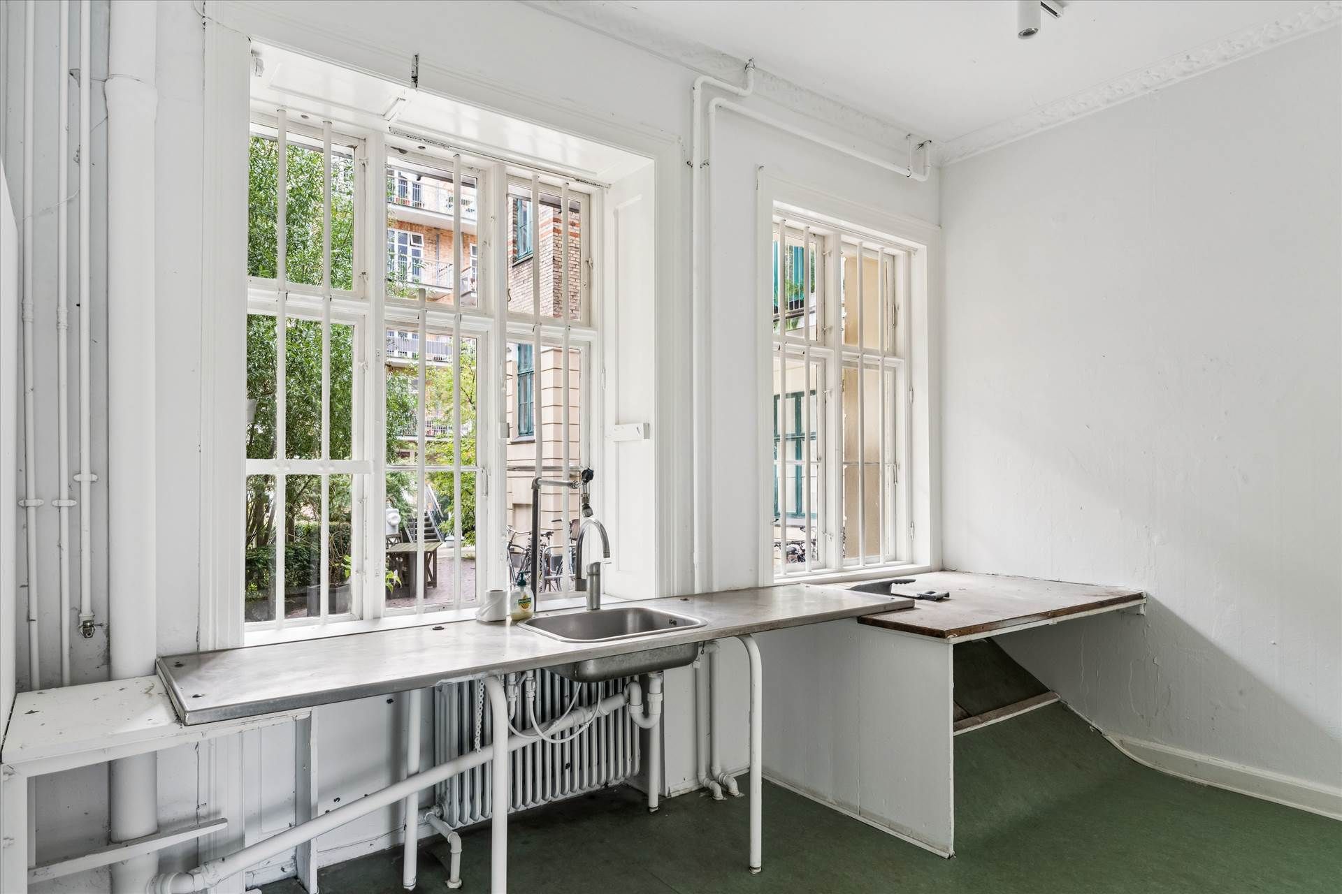 En hvidvaskede køkken med to vinduer og en rustfri stål skralvask samt et bord med et gammelt skriftstykke