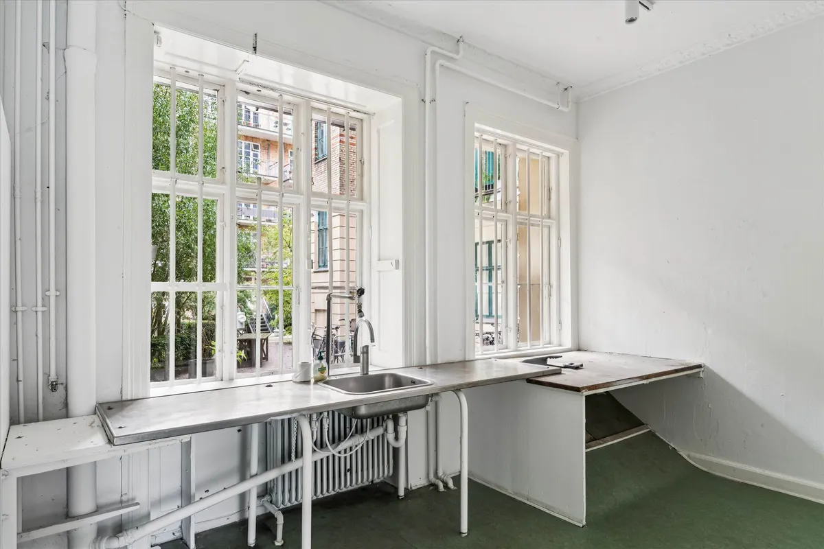 En hvidvaskede køkken med to vinduer og en rustfri stål skralvask samt et bord med et gammelt skriftstykke