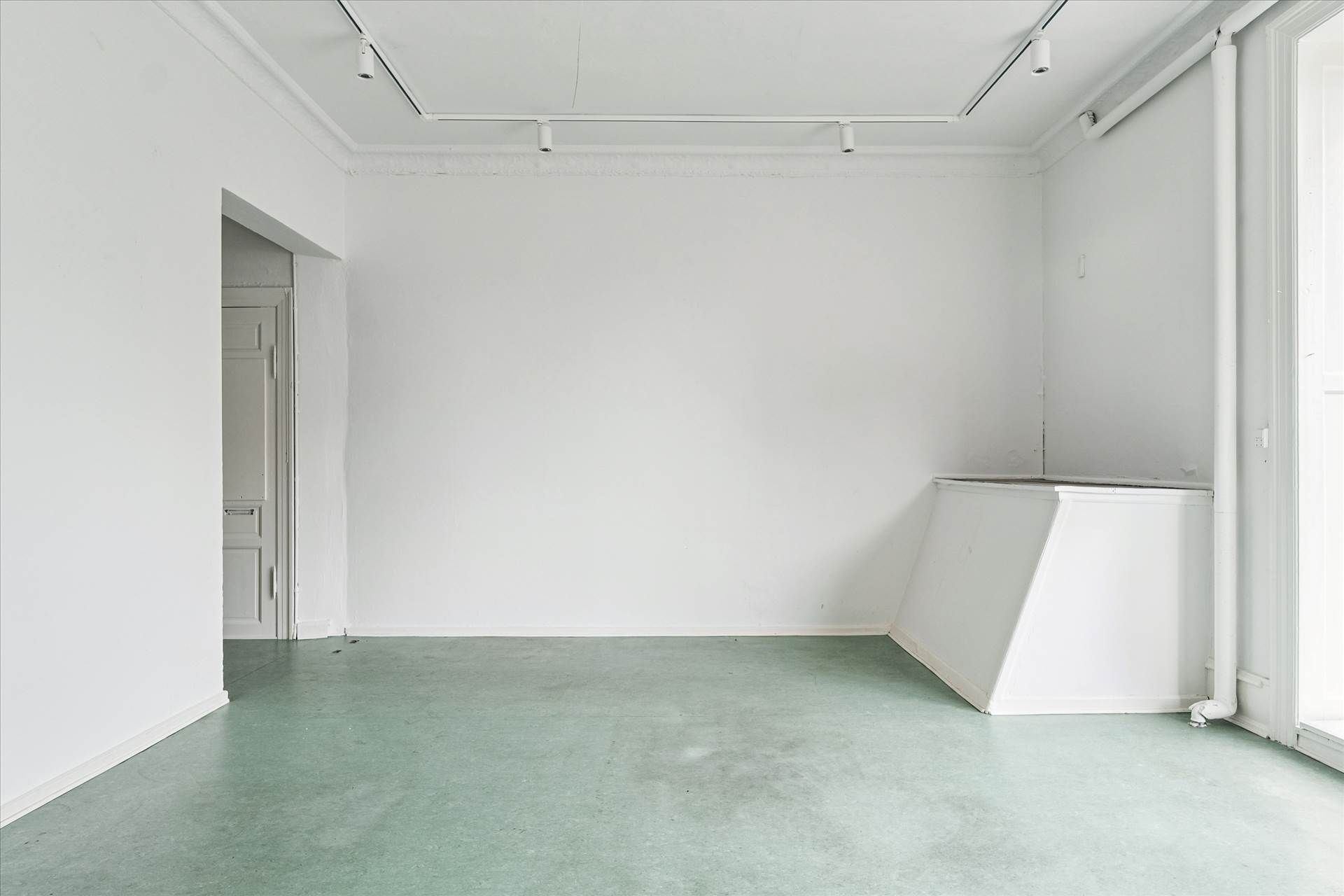 Et tomt rum med hvide vægge, grøn gulv og lyse armaturer i loftet. Et hjørne med dør og vindue.