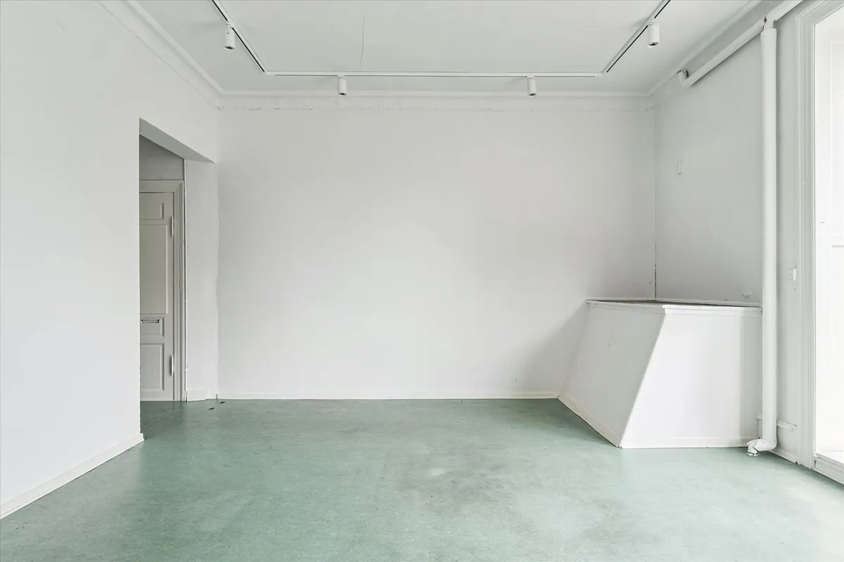 Et tomt rum med hvide vægge, grøn gulv og lyse armaturer i loftet. Et hjørne med dør og vindue.