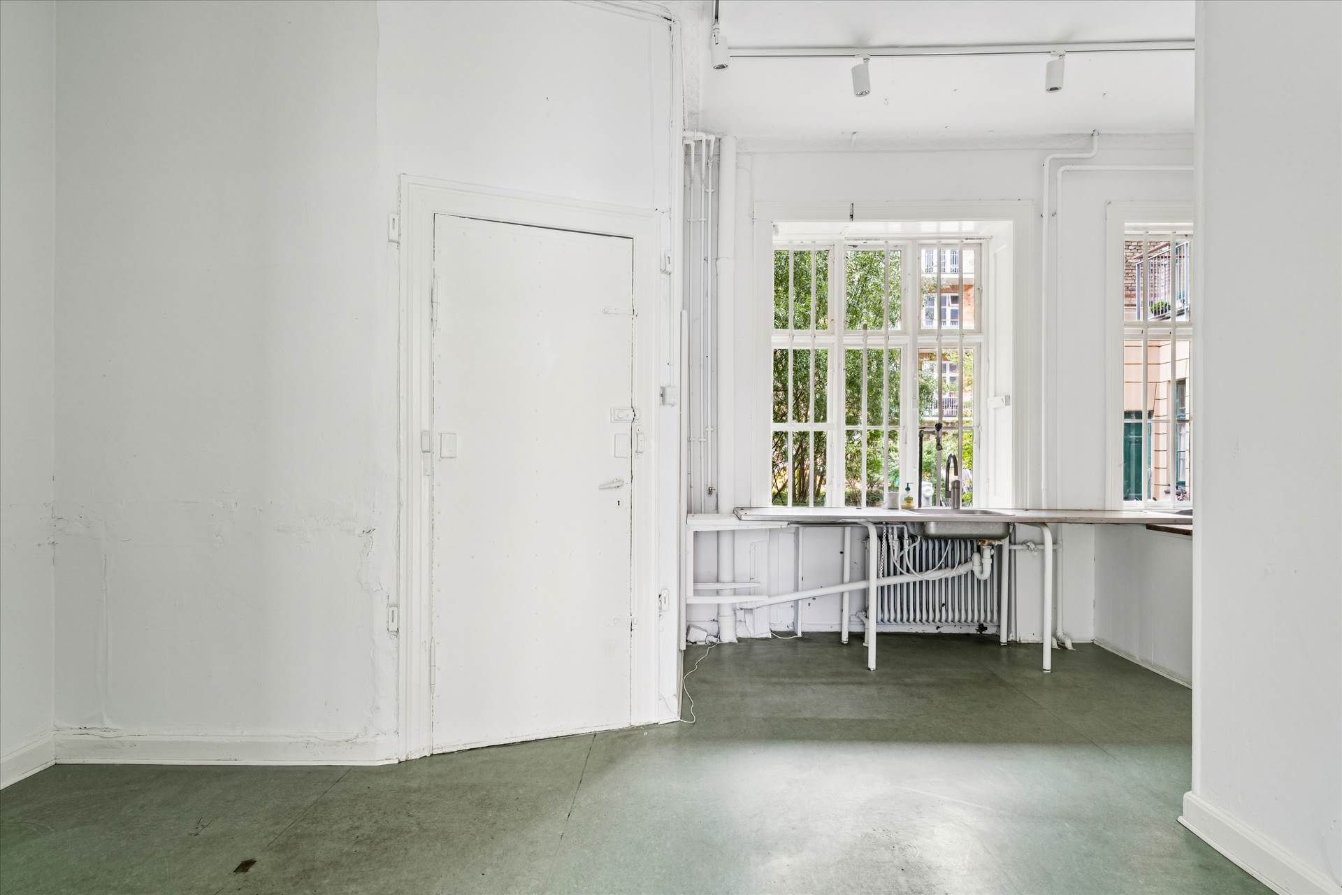 En tom hvid malet stue med lyse gulvbelægning og to vinduer. Der er en hvid dør med håndtag og to lysekroner i loftet.