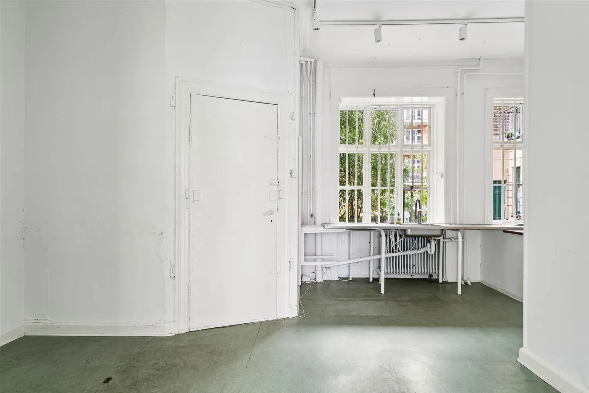 En tom hvid malet stue med lyse gulvbelægning og to vinduer. Der er en hvid dør med håndtag og to lysekroner i loftet.