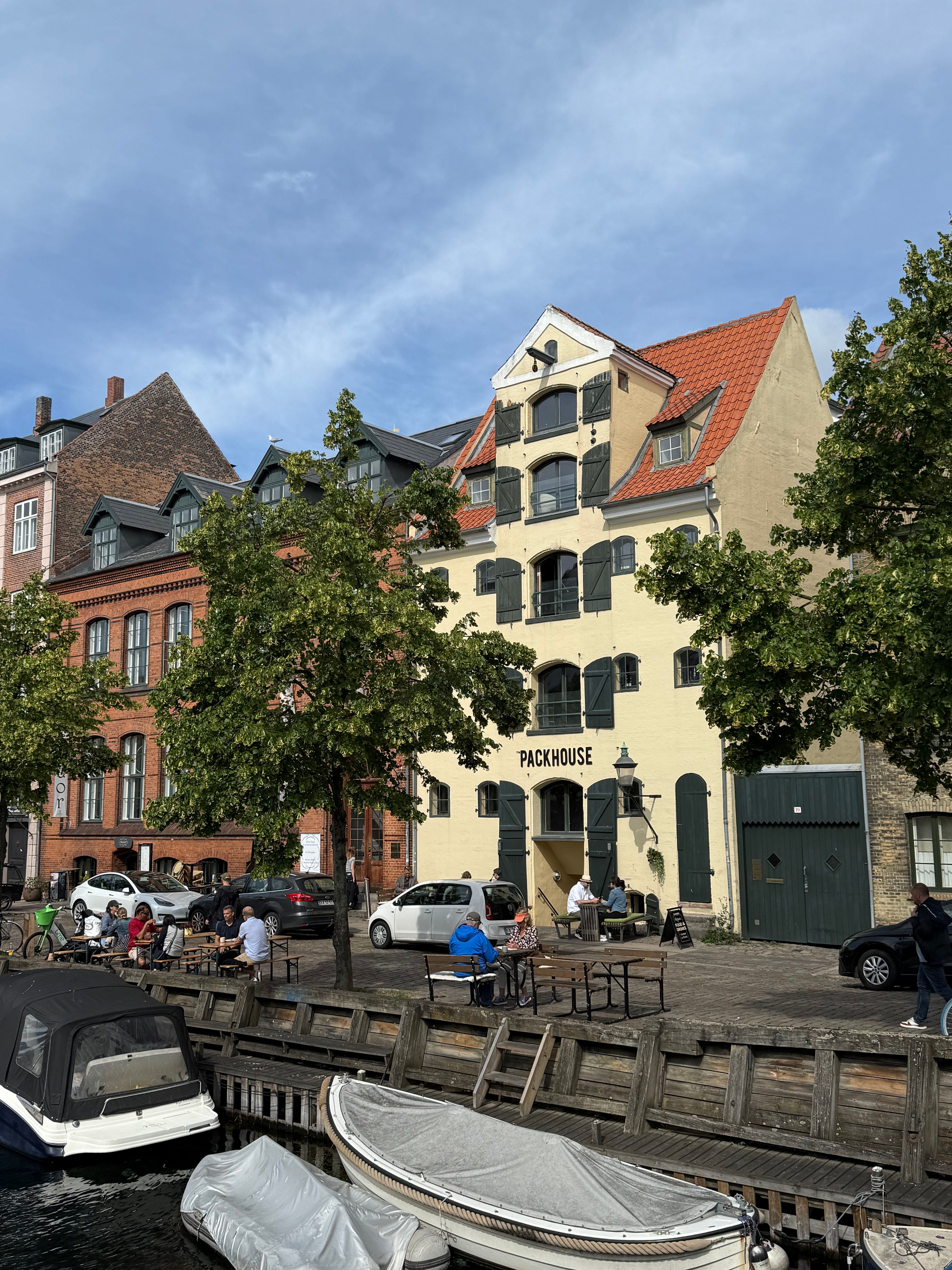 Mennesker sidder ved borde ved en havn med flere både og en bygning med navnet Packhouse
