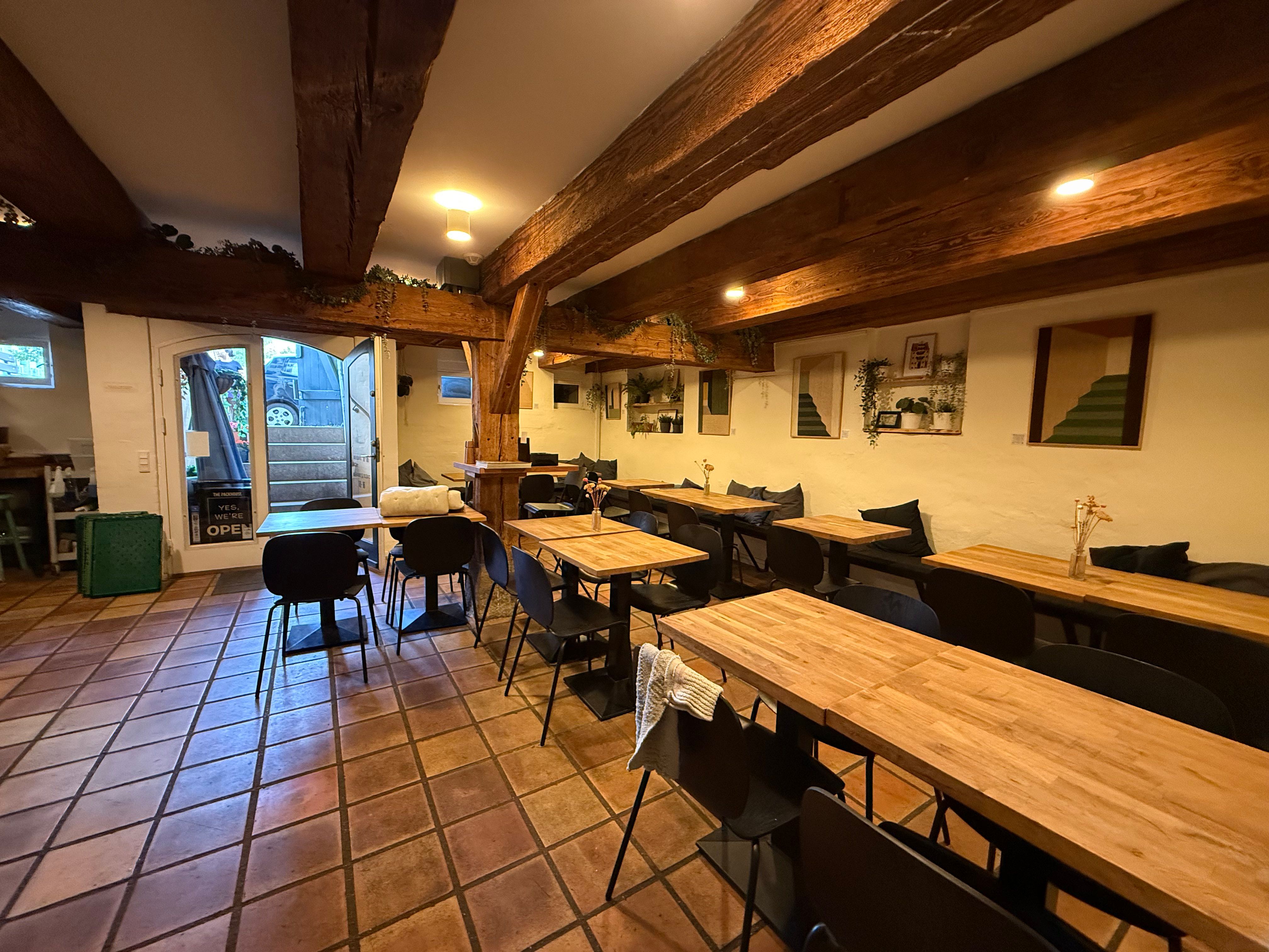 Et moderne restaurant med trægulve og bord, stole, plante og lysekroner på loftet. Et vindue med en åbning og en trappe med