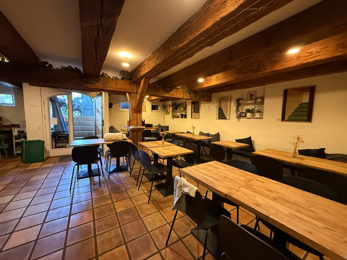 Et moderne restaurant med trægulve og bord, stole, plante og lysekroner på loftet. Et vindue med en åbning og en trappe med