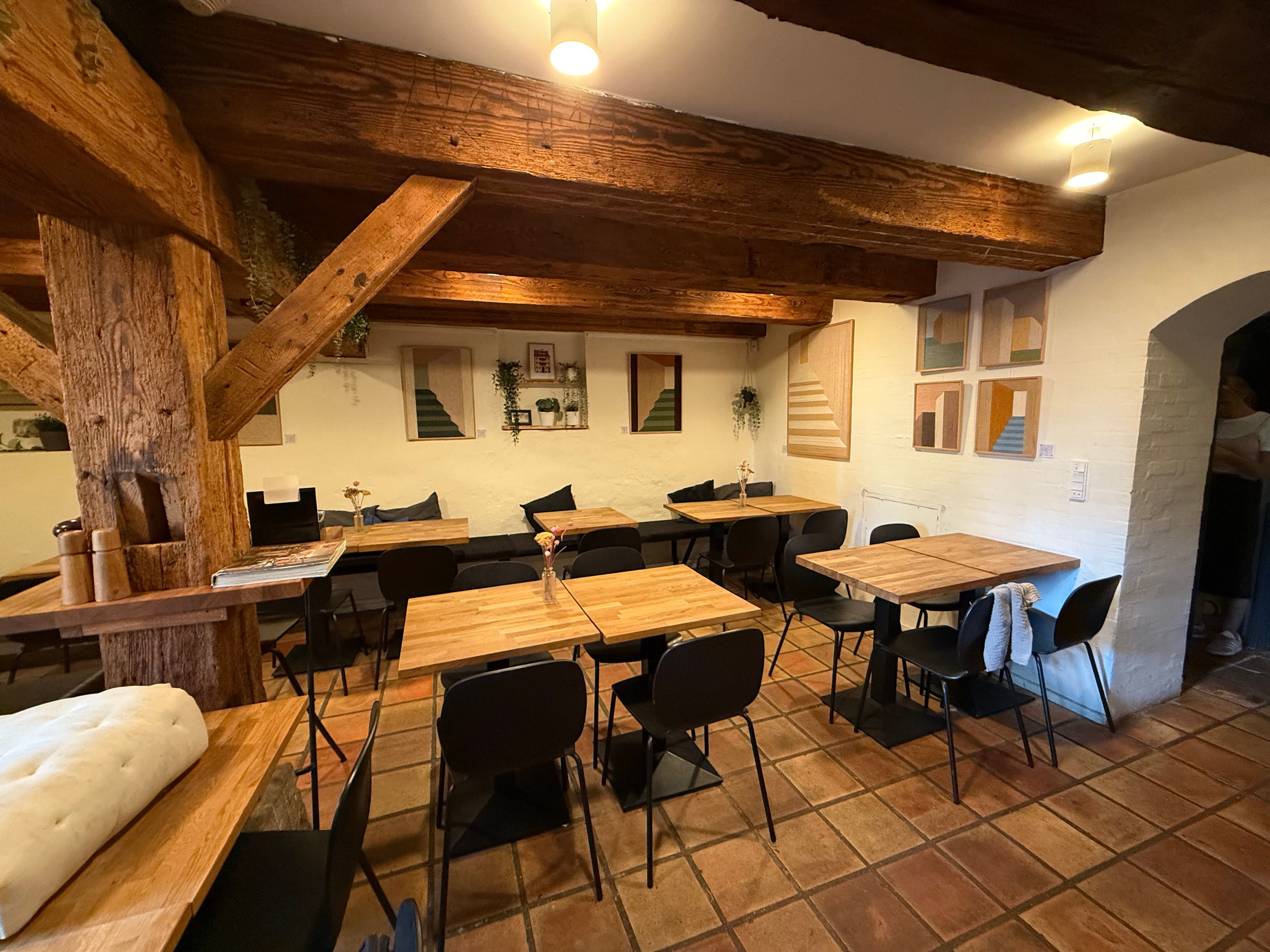 Lyserøde fliser og høje træbjælker, borde og stole, lysekroner og plante i lysekroner, malerier og stole på hver side af