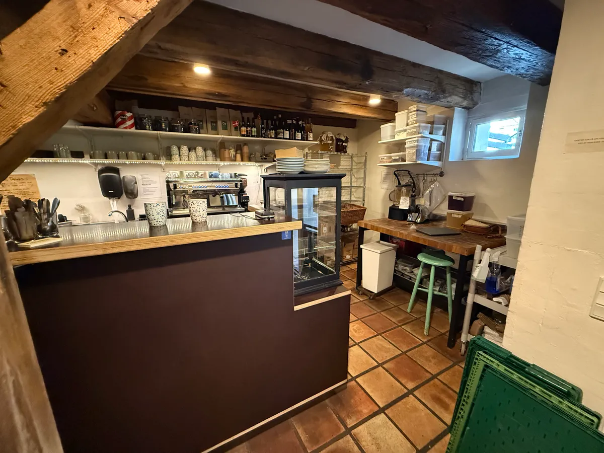 Et lille caférum med loftsbjælker, en bar, et kaffemaskin, en skuffe og en stol samt diverse udstyr på hylder