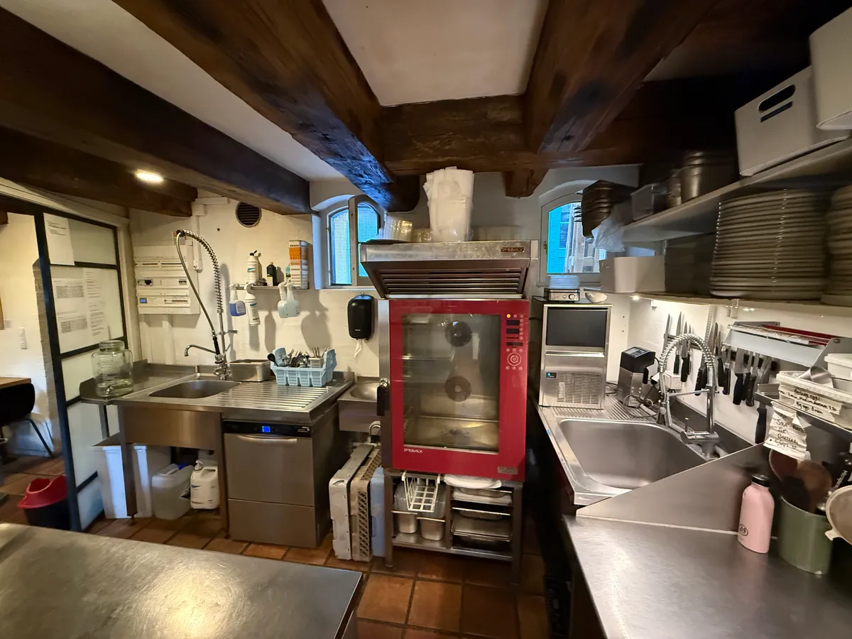 Køkken med rødt komfurapparat, flere stålskåle og glasflasker samt en stålbord og to lyse brædder