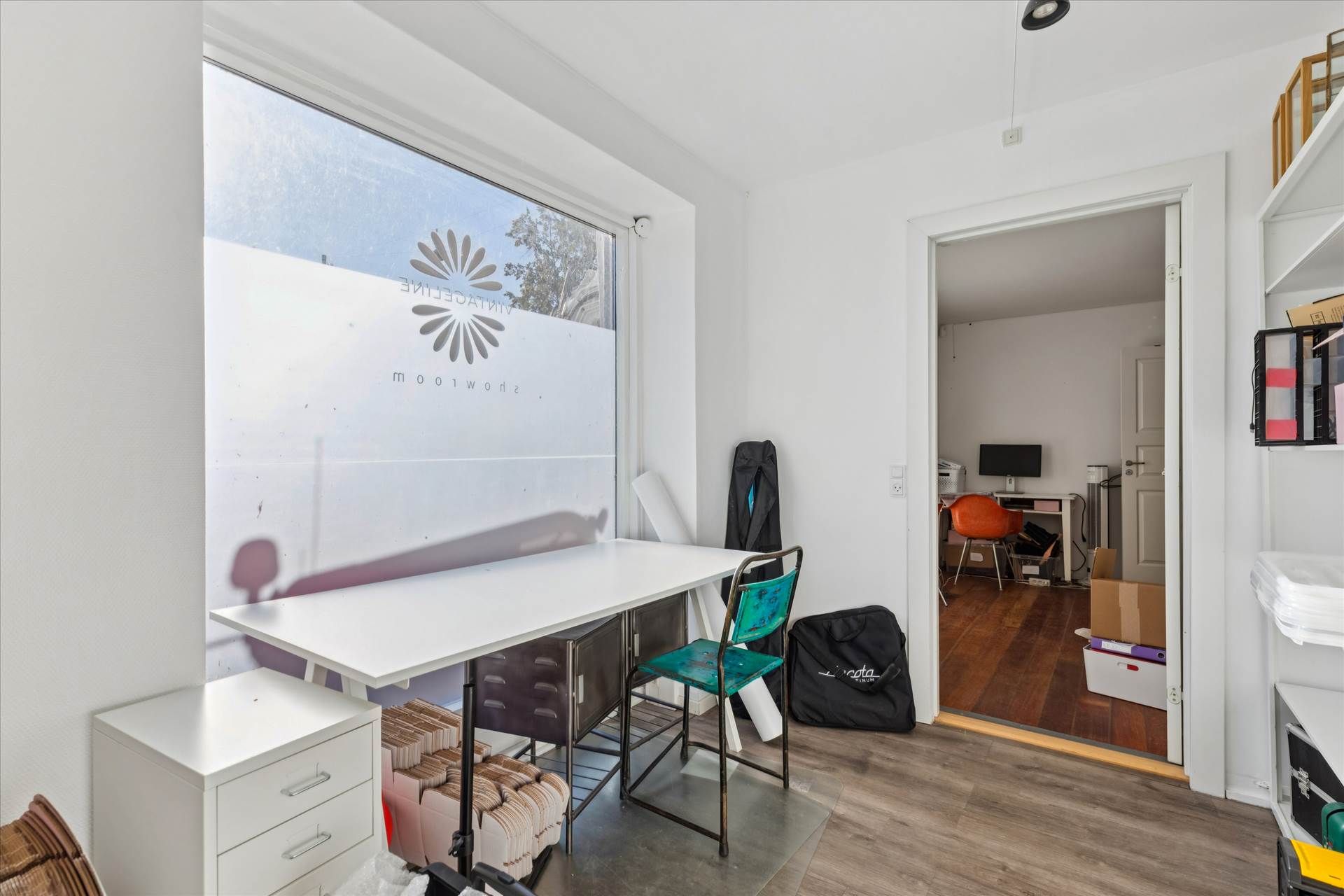 Studie med højloft, hvidt bord, to stole, stol med blåt sæde og en sort taske på gulvet